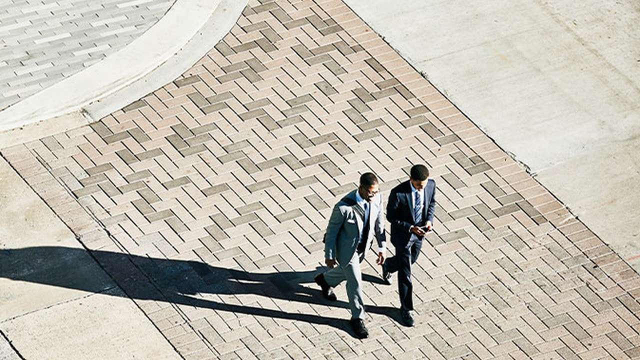 To mænd i jakkesæt går på et fortov, set oppefra.