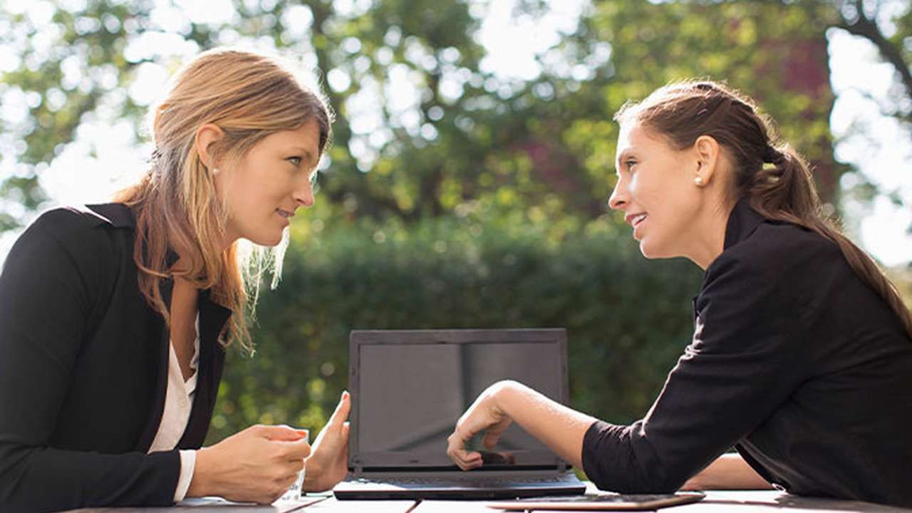 To kvinder sidder ved et bord udendørs og arbejder på en bærbar computer.