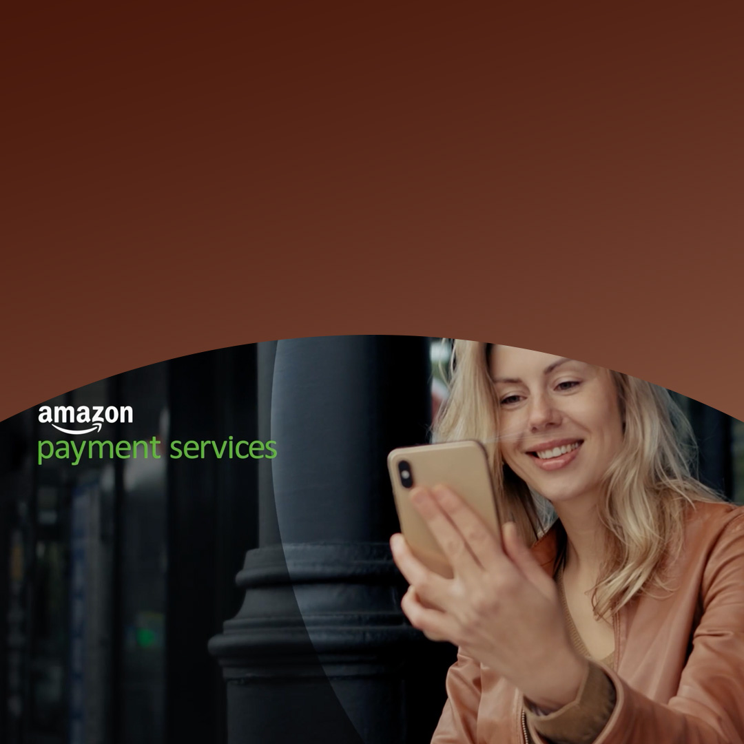 Kvinde i en brun jakke, der smiler til sin smartphone, mens hun bruger Amazon-betalingstjenester med Mastercard-gateway.