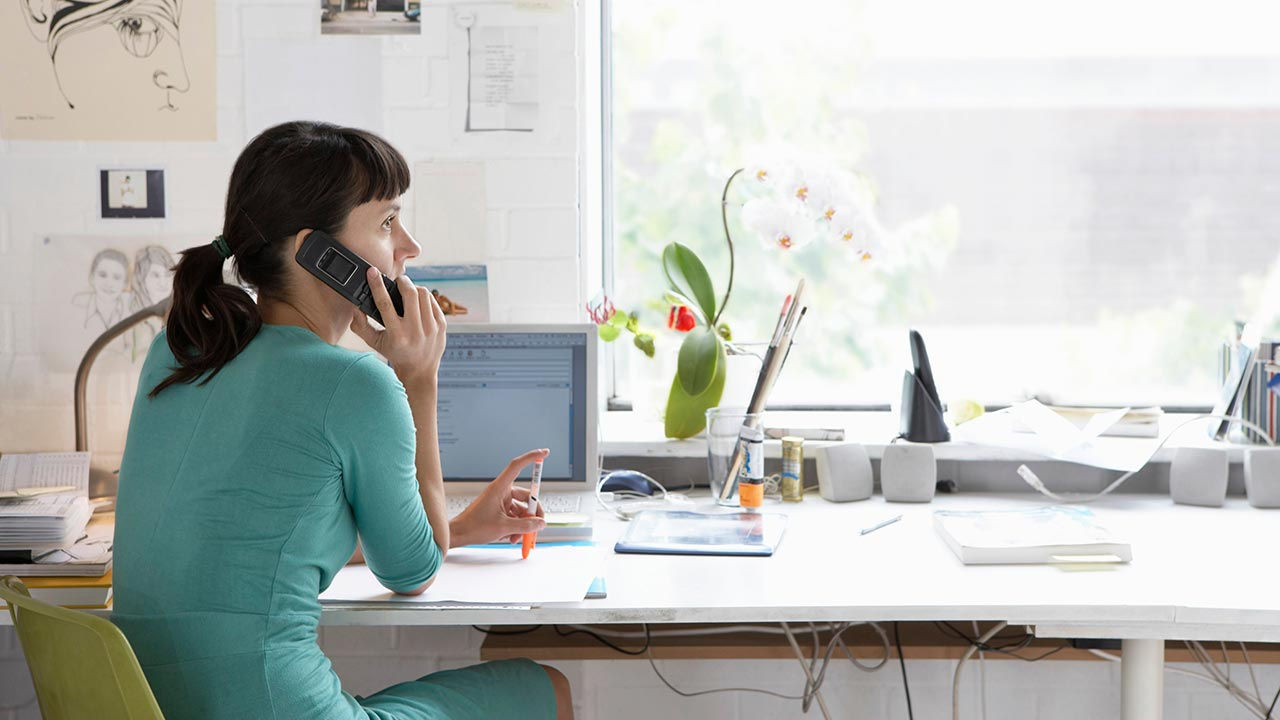 En kreativ kvinde sidder ved et skrivebord på sit hjemmekontor og taler i en trådløs telefon, mens hun holder en orange tusch i den anden hånd.