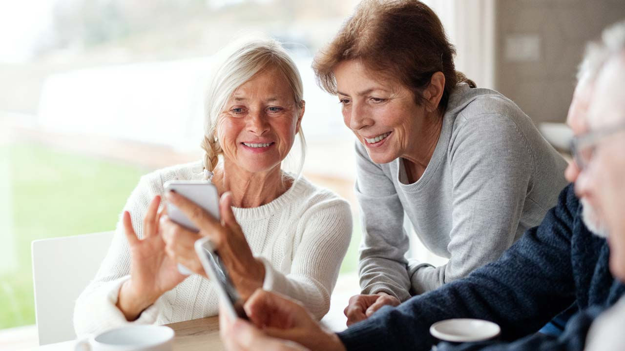 To personer sidder ved et bord og ser på en smartphone, mens en tredje person holder en tablet. Der står kaffekopper på bordet, og baggrunden viser et lyst rum med store vinduer.