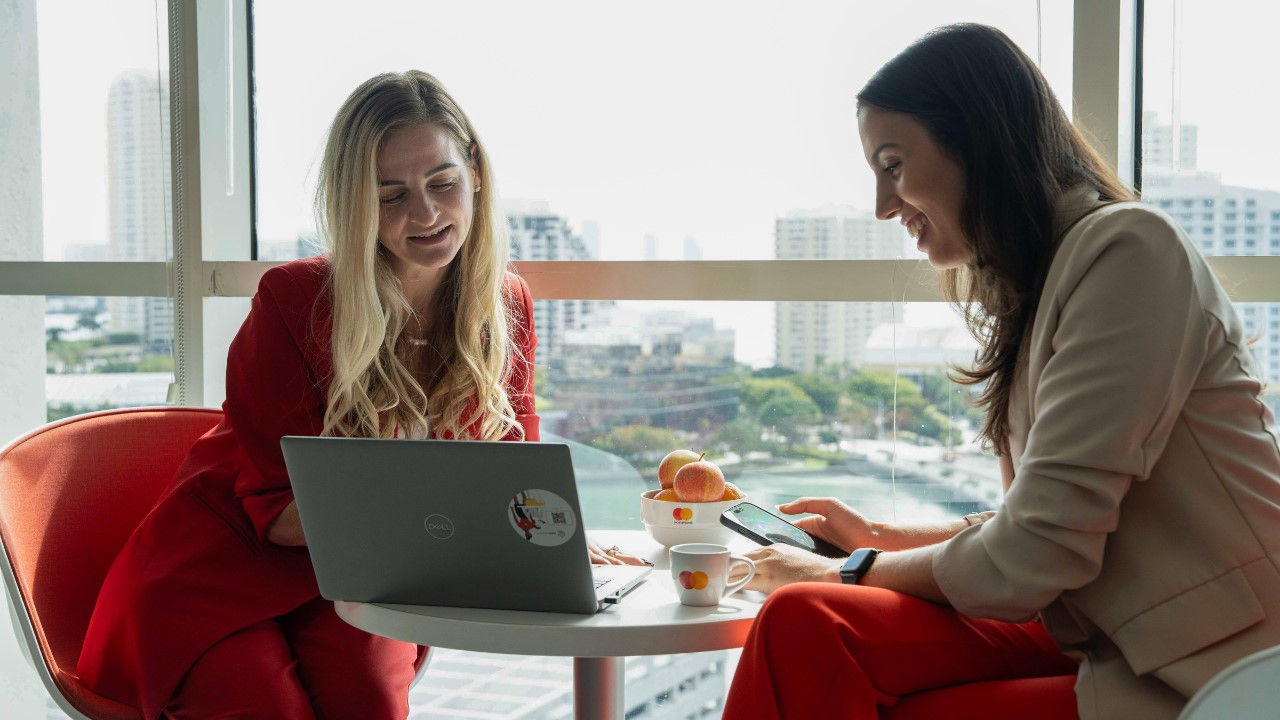 Two employees at Mastercard's Miami office confer over a computer in front of a large window overlooking the skyline. 