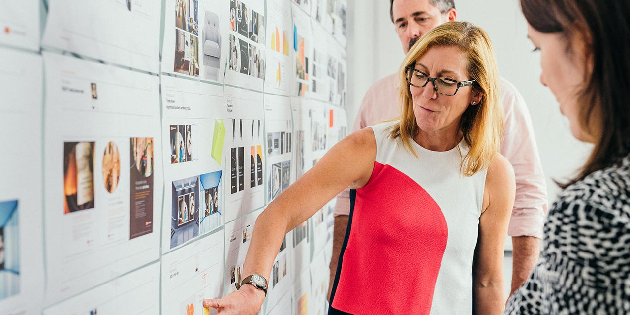 Woman presenting ideas on a wall of design mockups while a colleague stands behind her.