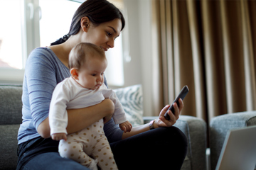 Mother and baby sitting on a couch together, both focused on a smartphone screen.