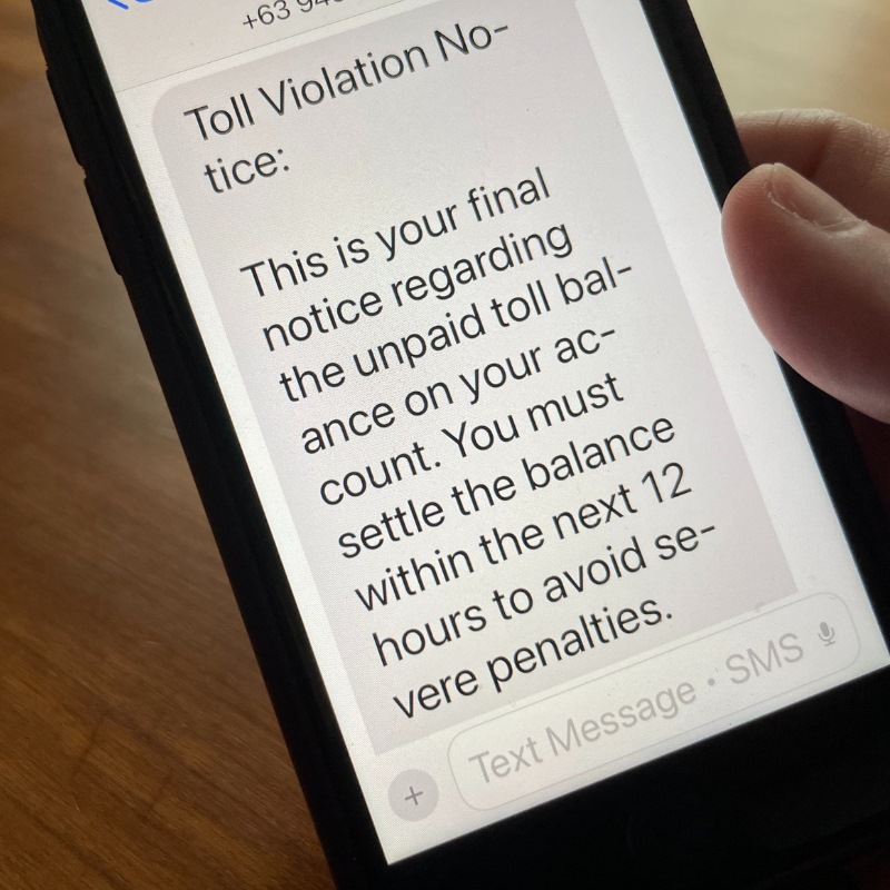 A person holds a cell phone with a text scam notification on it about a toll violation.
