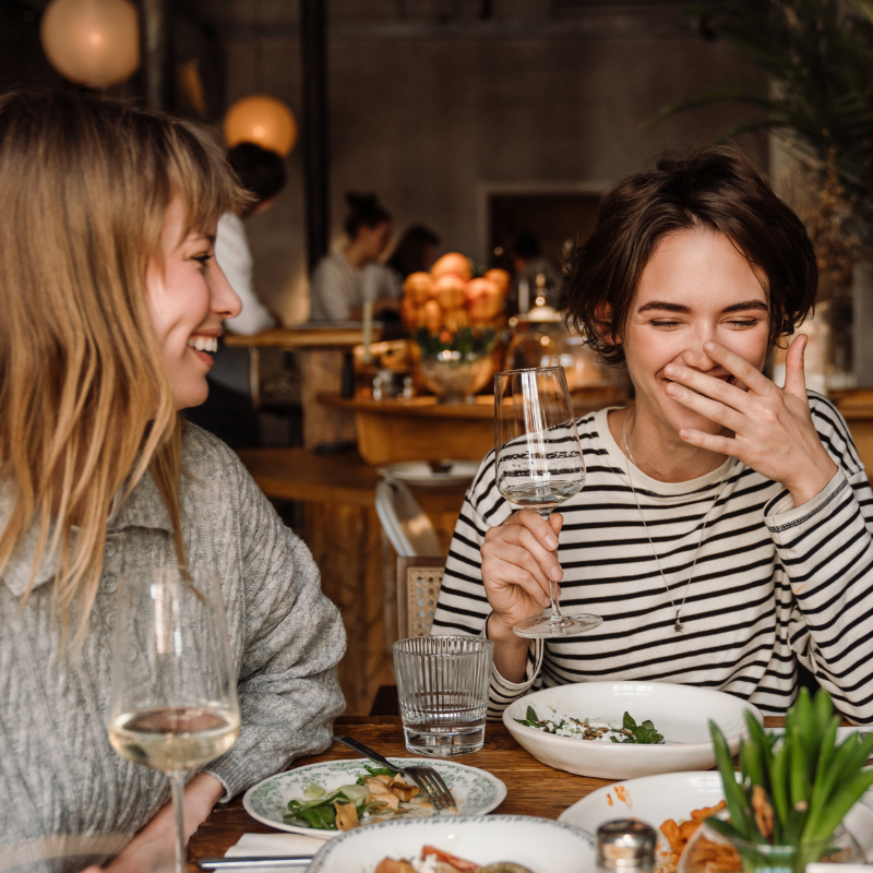 A woman holds a wine glass and laughs while a friend looks on in a restaurant. 