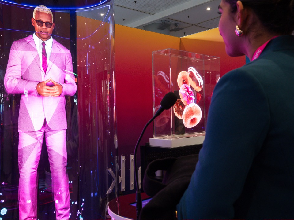 An NRF attendee stands in front of a microphone in front of a hologram of a stylishly-dressed man in sunglasses. 