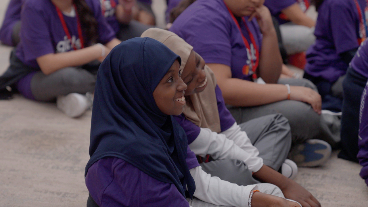 Kaksi henkilöä istuu maassa kuunnellen esitystä.