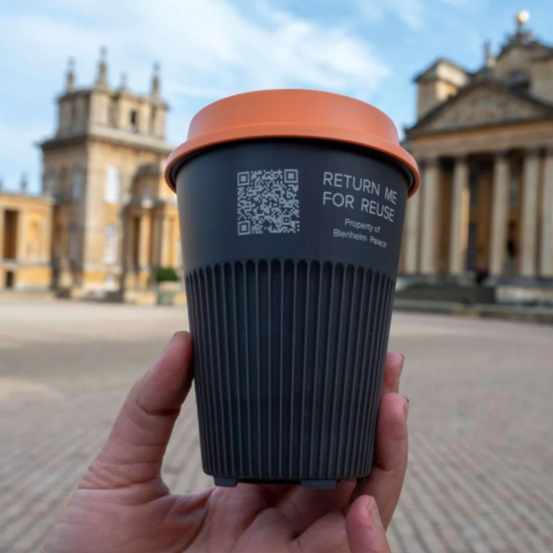 A reusable cup with a QR code for return is held in front of Blenheim Palace. 