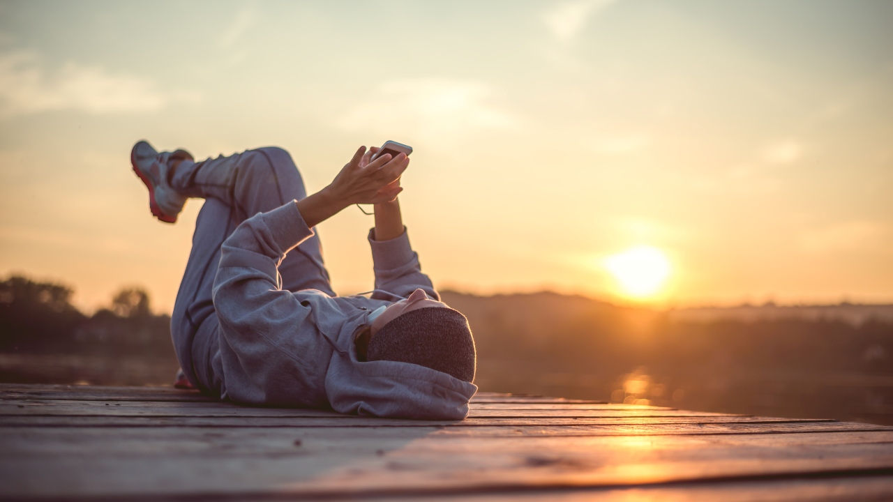 A woman lays on a dock in a running gear holding her phone as the sun rises. 