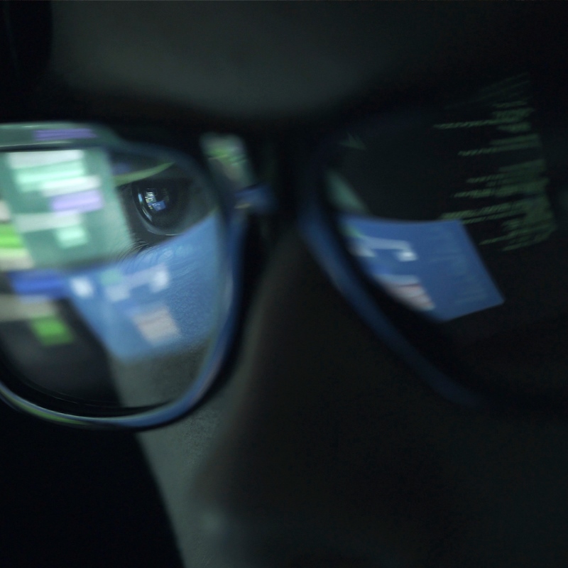 A man in a dark room with computer coding reflected in his eyeglasses. 