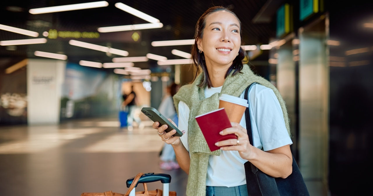 A woman holds a smartphone, passport and coffee cup in her hand with a suitcase next to her in a transit hub. 