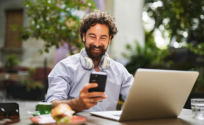 Smiling man holding a smartphone while sitting outdoors with a laptop.