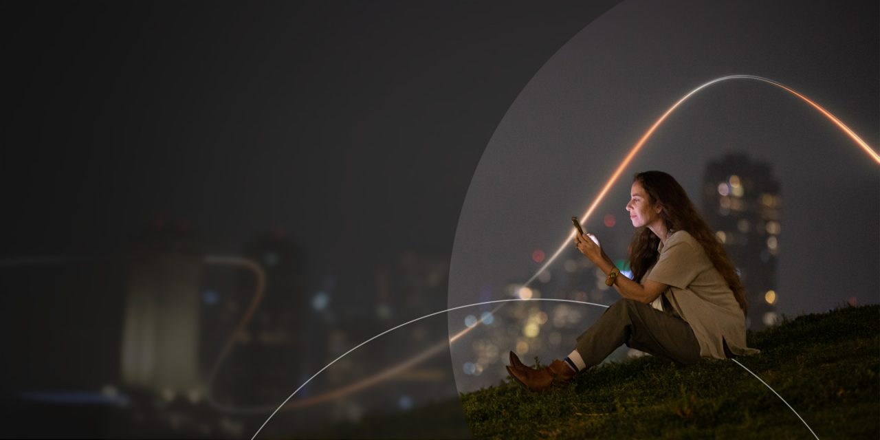 Woman sitting on grass at night, using her phone, with blurred city lights glowing in the background.