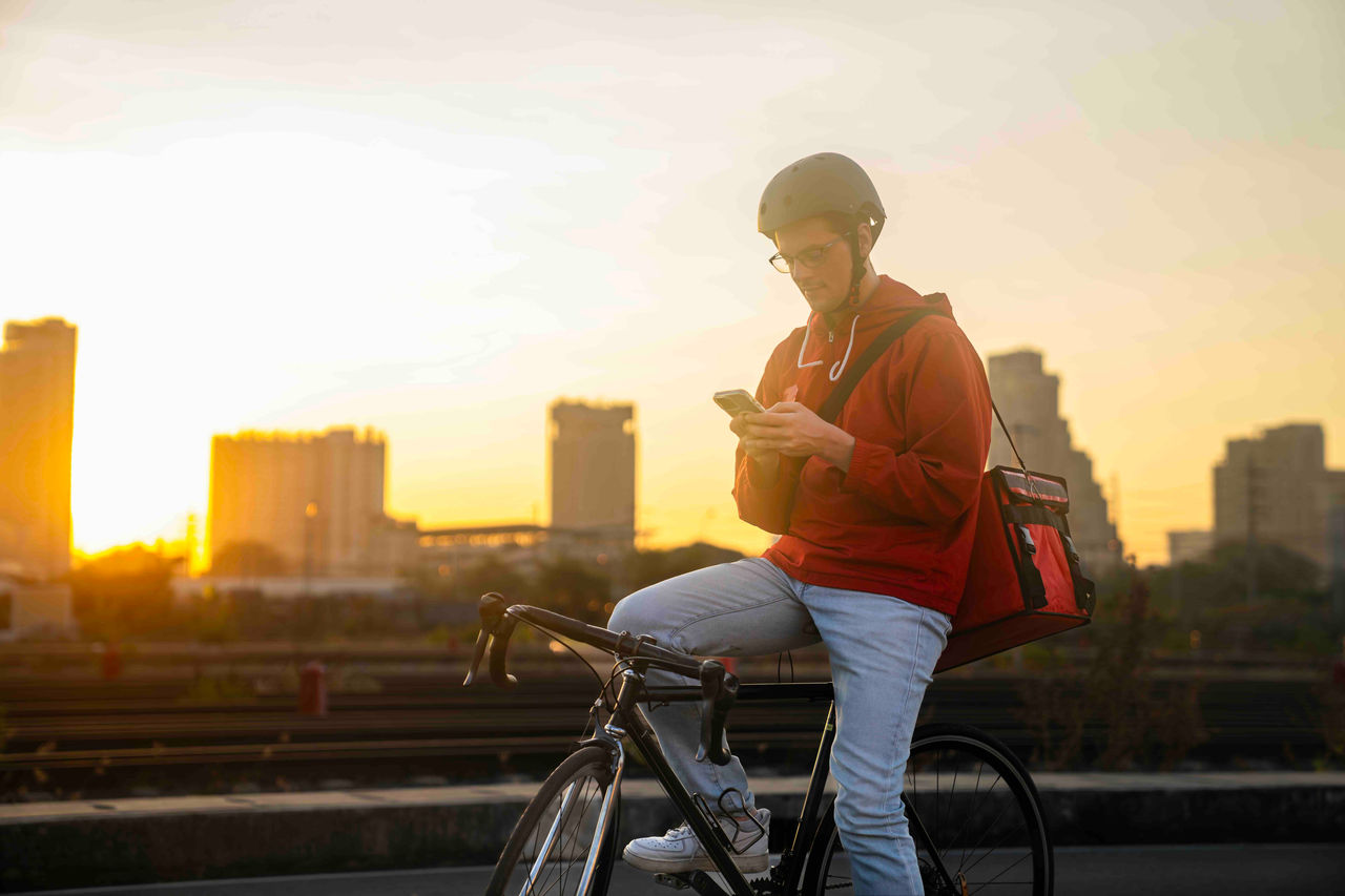Gig worker looking at phone while sitting on bike at sunset