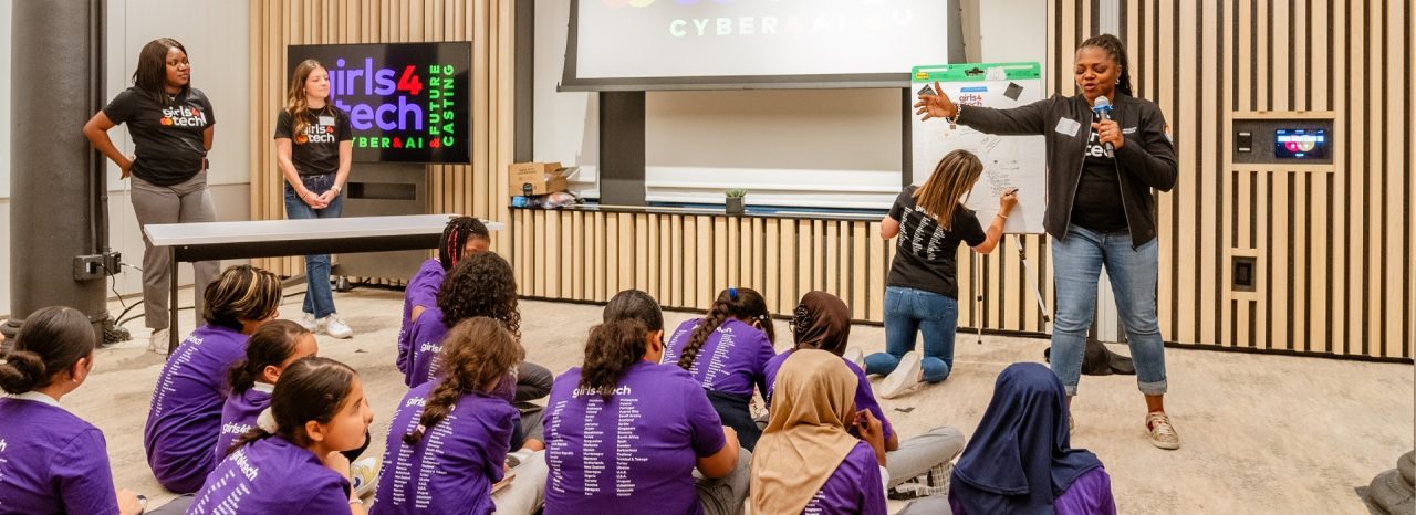 Alissa "Dr Jay" Abdullah stands in front of a group of young female students in purple Girls4Tech T-shirts, seated on the floor, to discuss future cyber risks.