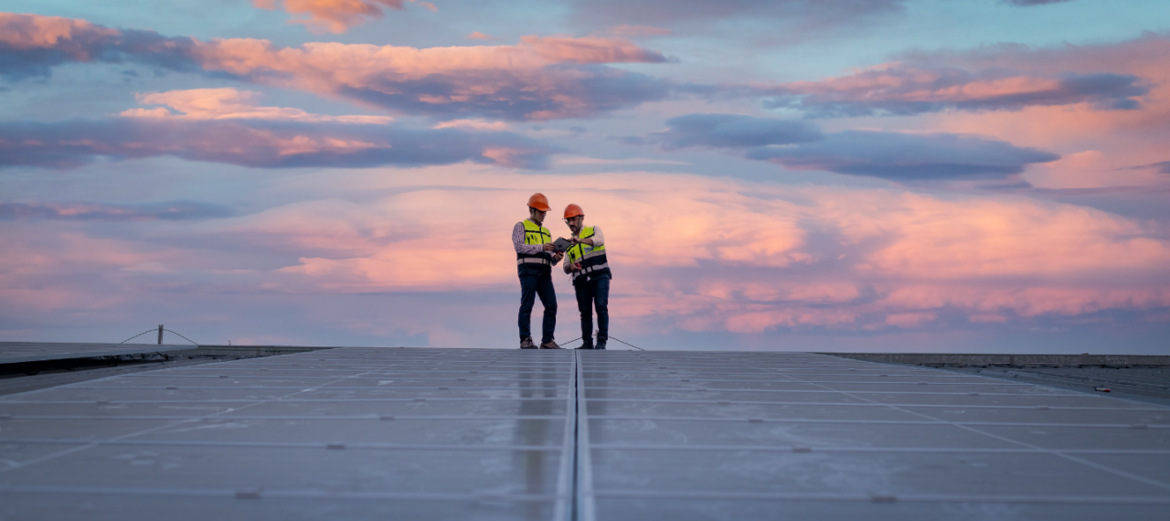 Two men in work vests stand on top of a field of solar panels at sunrise. 