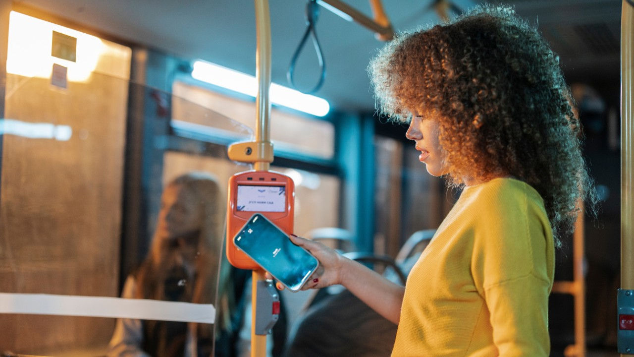 A woman holds a phone up to a card reader in a bus. 