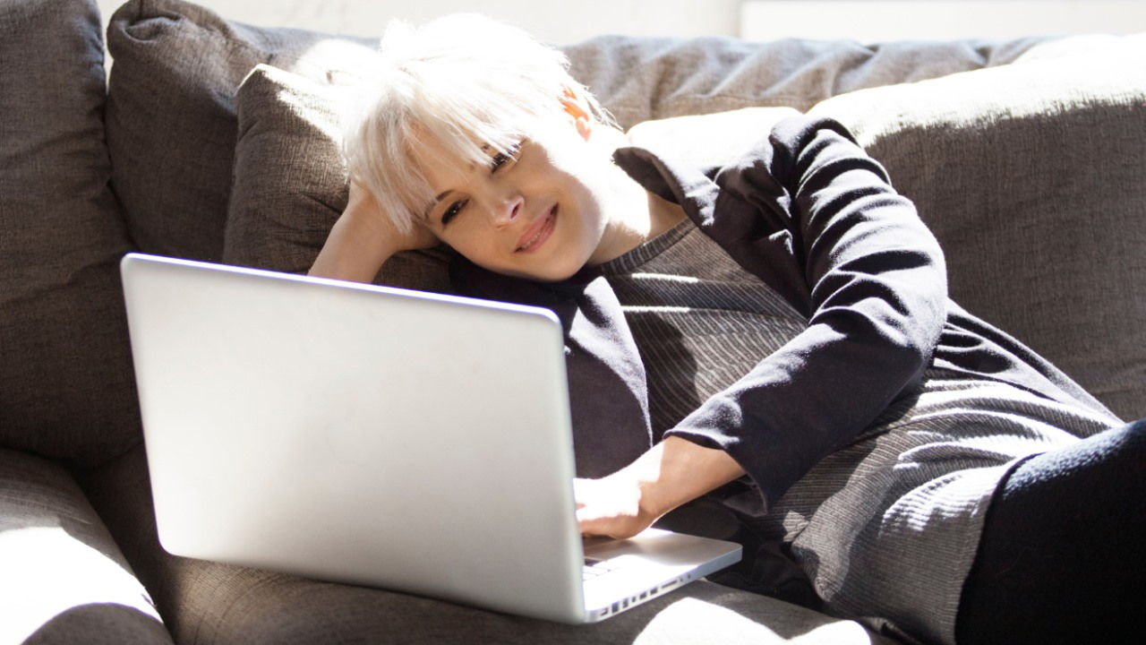 A woman lays on a couch smiling at her laptop. 