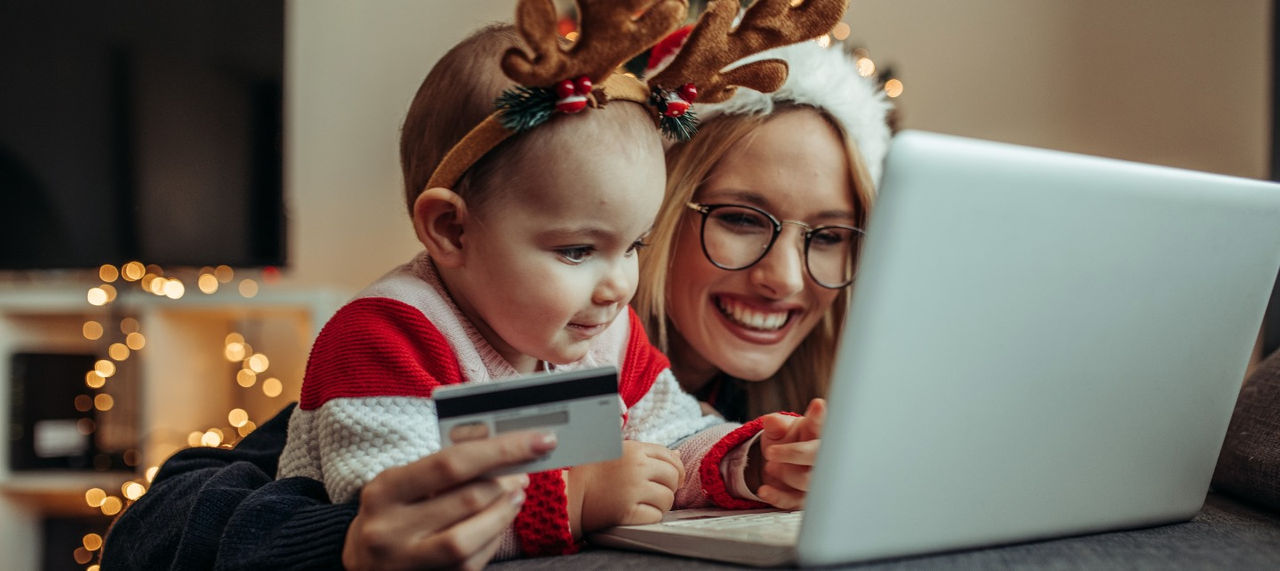 A woman in a Santa hat and a young girl in an antler headband look at a laptop while the woman holds a credit card. 