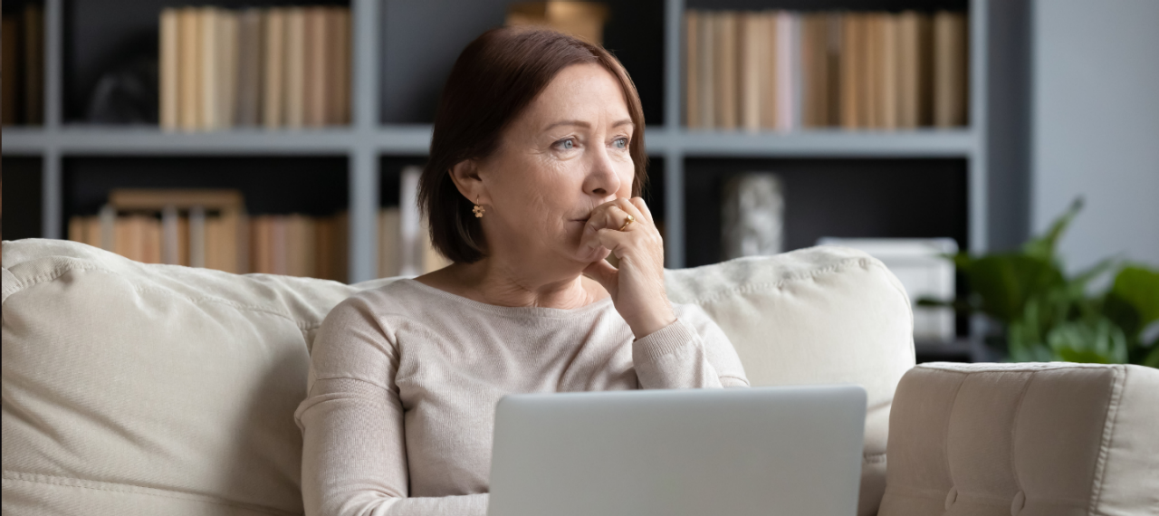 A middle-aged woman looks concerned  as she stares into the distance with a laptop on her lap while sitting on a couch. 