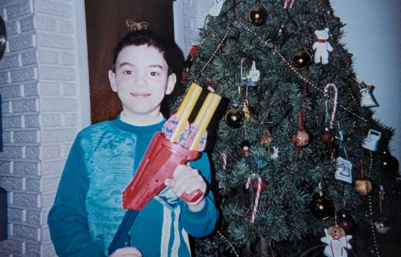 Chris Mullen as a child in the 1990s in front of a christmas tree holding a toy. 