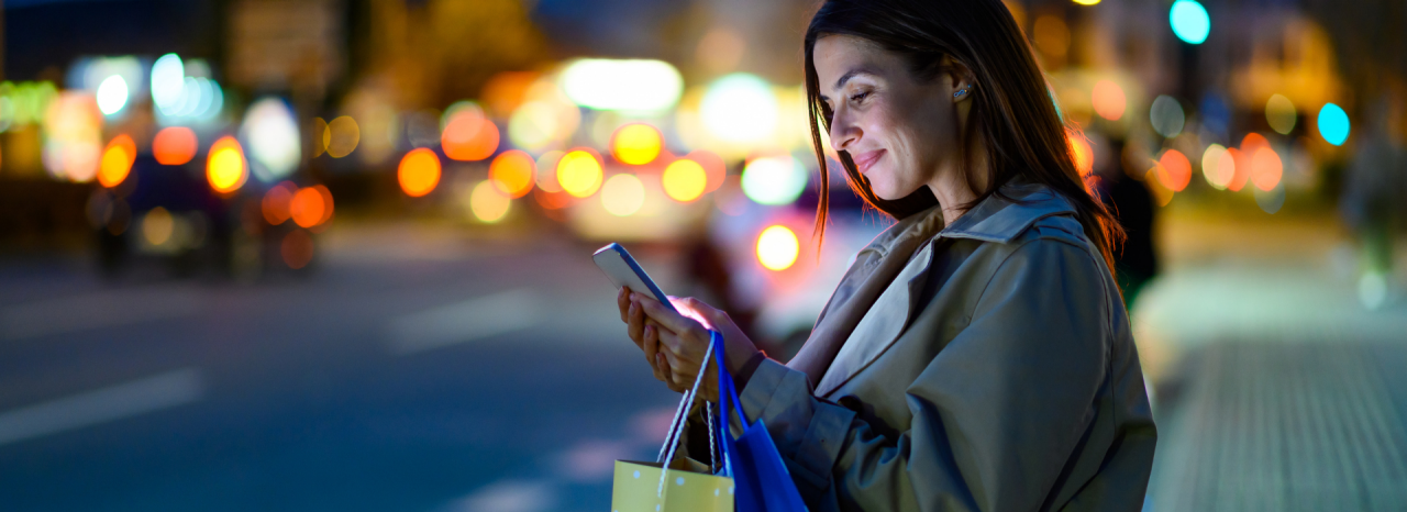 買い物袋を持った女性が、人通りの多い通りの近くでスマートフォンを見ている。 