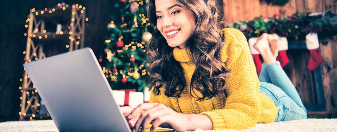 黄色いセーターを着た女性が、クリスマスツリーの前で床に横たわり、ノートパソコンを見ている。 