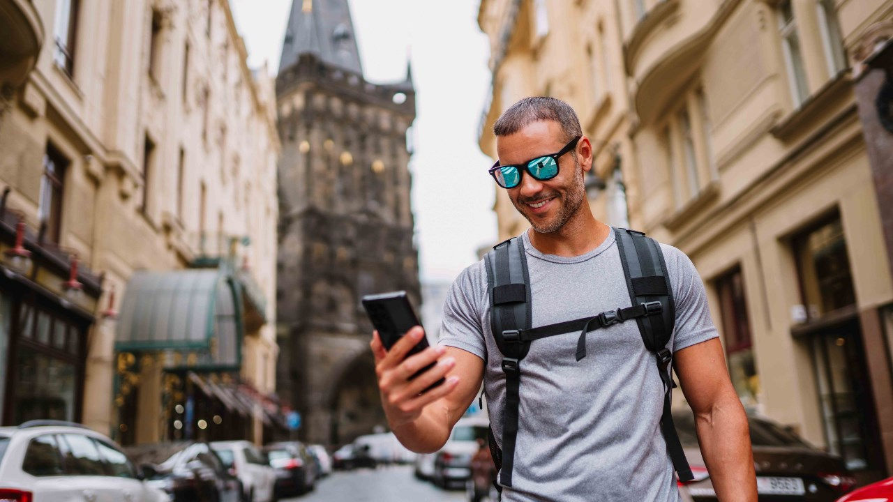 A traveler wearing sunglasses and a backpack smiles at his phone in a city. 