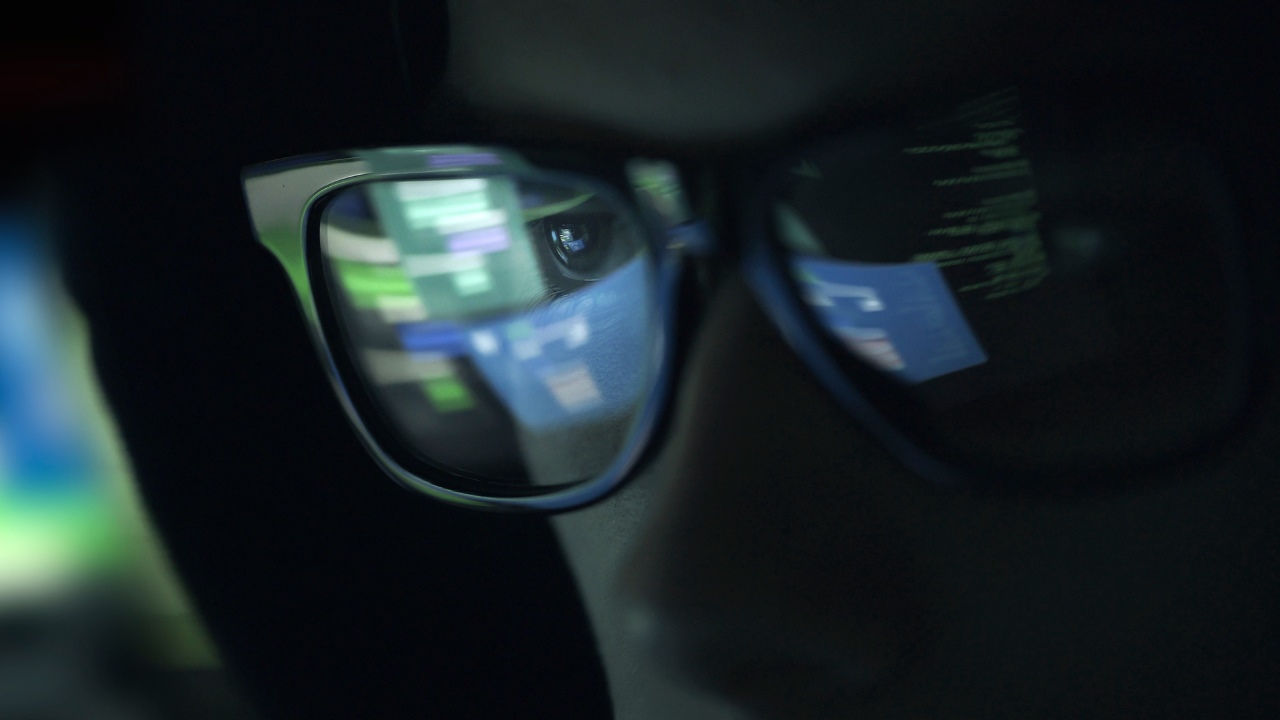A man in a dark room with computer coding reflected in his eyeglasses. 