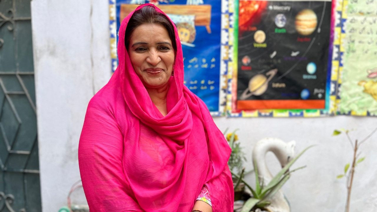 A woman in a pink shawl stands in front of a school, smiling. 