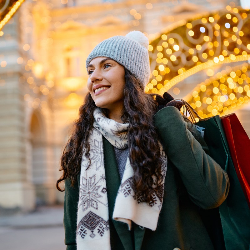 A woman dressed in a winter hat and scarf walks beneath arches covered with holiday lights while she carries shopping bags. 