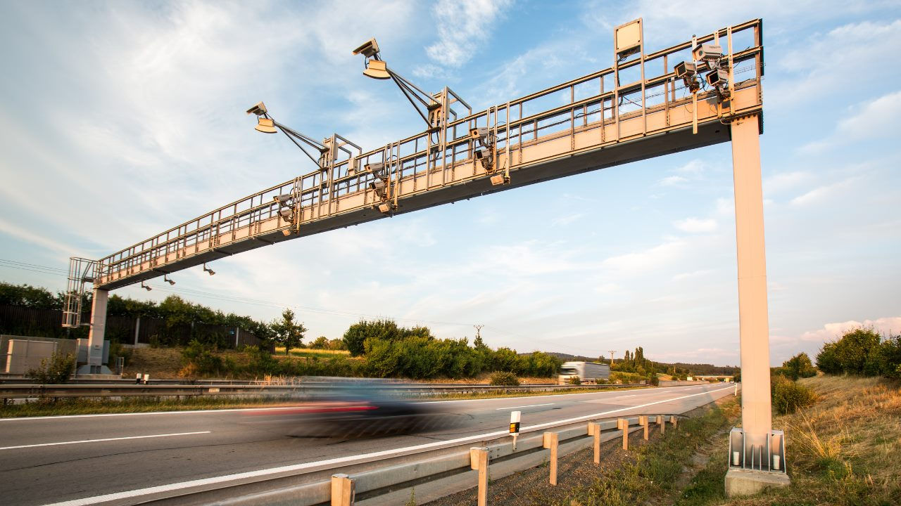 A car speeds beneath a toll gantry equipped with cameras on a highway. 