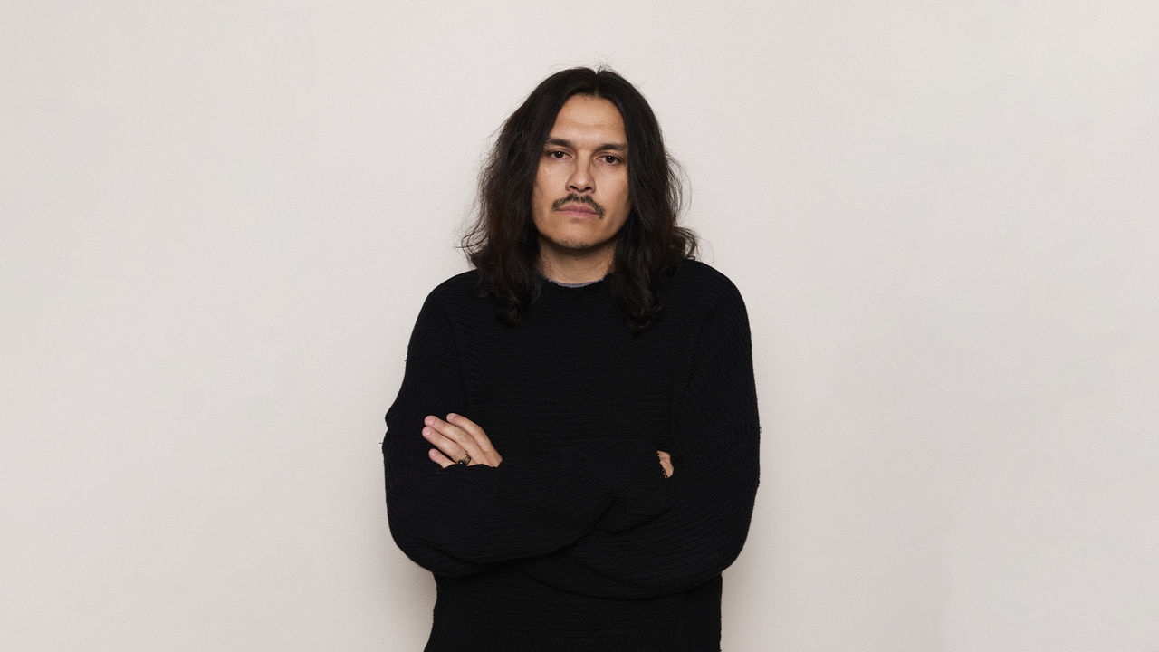 Portrait of fashion designer Guillermo Andrade standing with arms crossed, wearing a black textured sweater, against a plain off-white background.