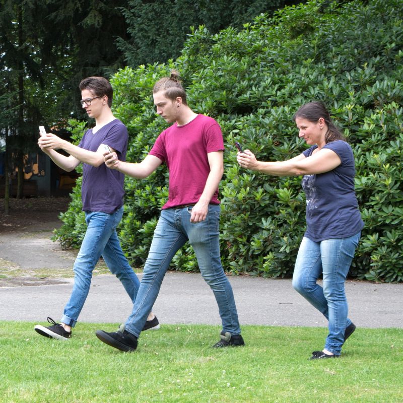 Three young people in jeans and t-shirts walk across a park holding their phones. 