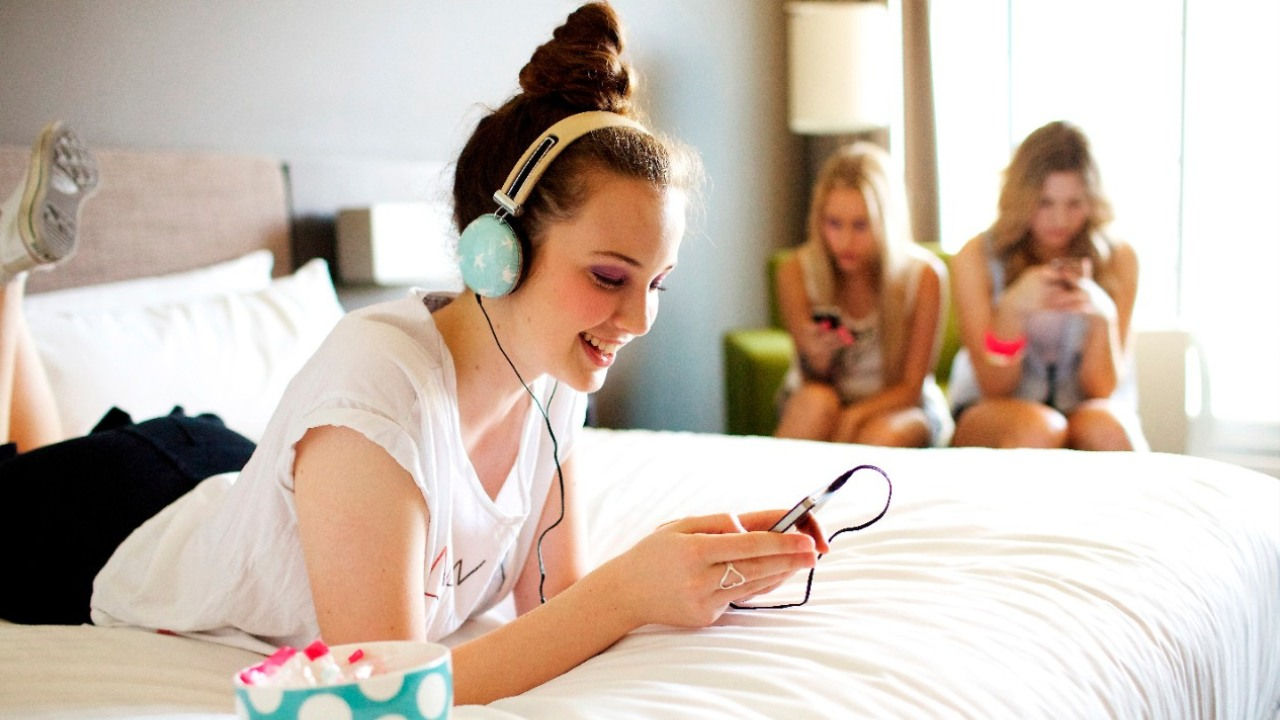 A girl wearing headphones look at her phone on her bed while two girls sit nearby also looking at their phones. 