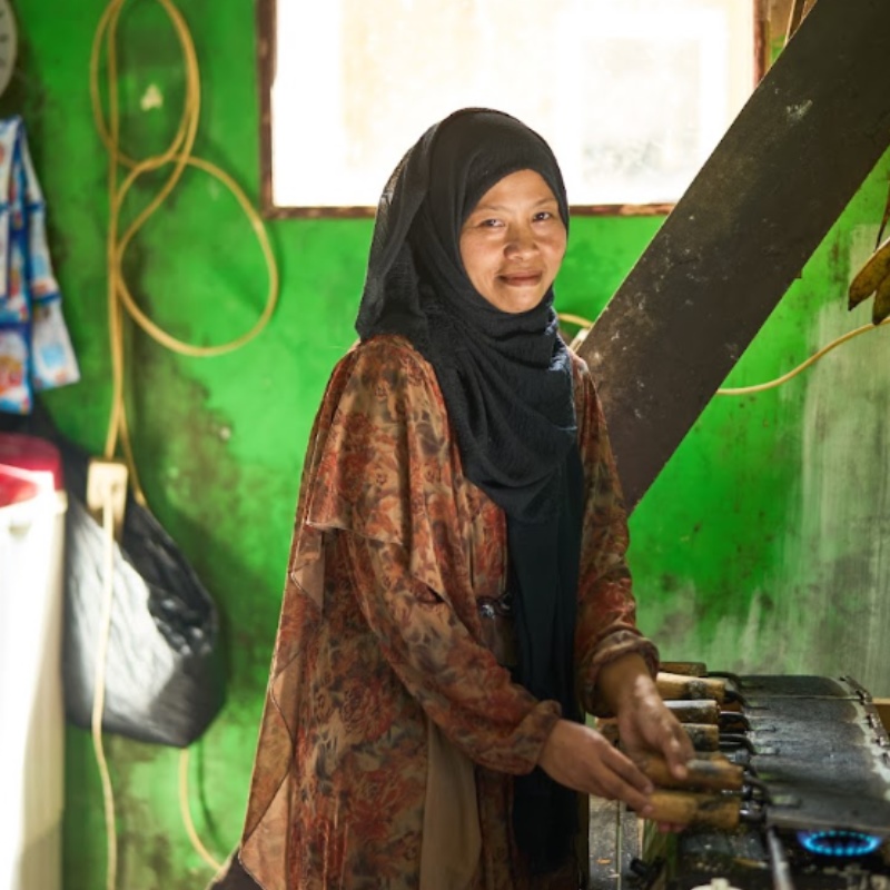 A woman in a headscarf stands in her kitchen over a stove. 