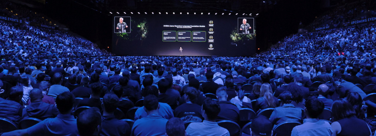 قاعة مؤتمرات مضاءة باللون الأرجواني مليئة بالناس مع مسرح في الطرف البعيد. 