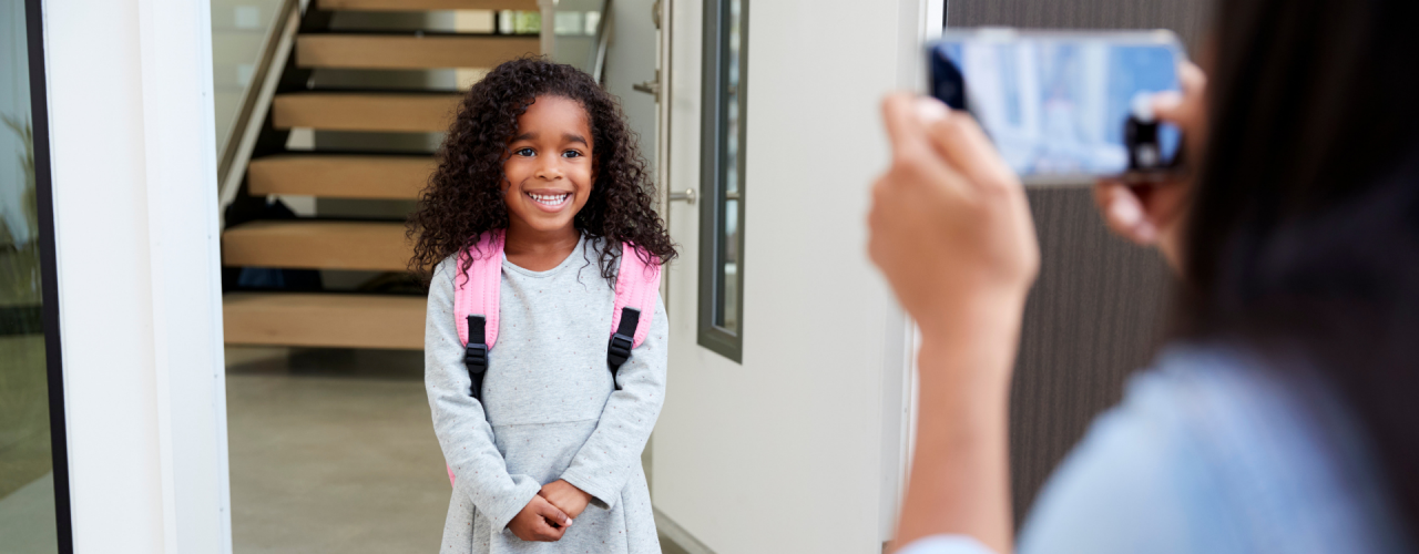 Une jeune fille portant un sac à dos pose pour la photo de son premier jour d'école devant sa mère, dans l'embrasure de la porte.