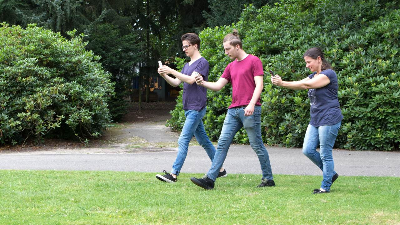 Three young people in jeans and t-shirts walk across a park holding their phones. 