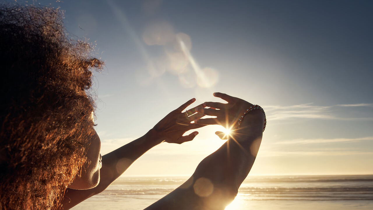 Se ve a una mujer tocándose la mano mientras disfruta de la luz del sol. La escena captura la belleza de la naturaleza con un cielo despejado, nubes y un entorno de playa sereno durante la puesta de sol.