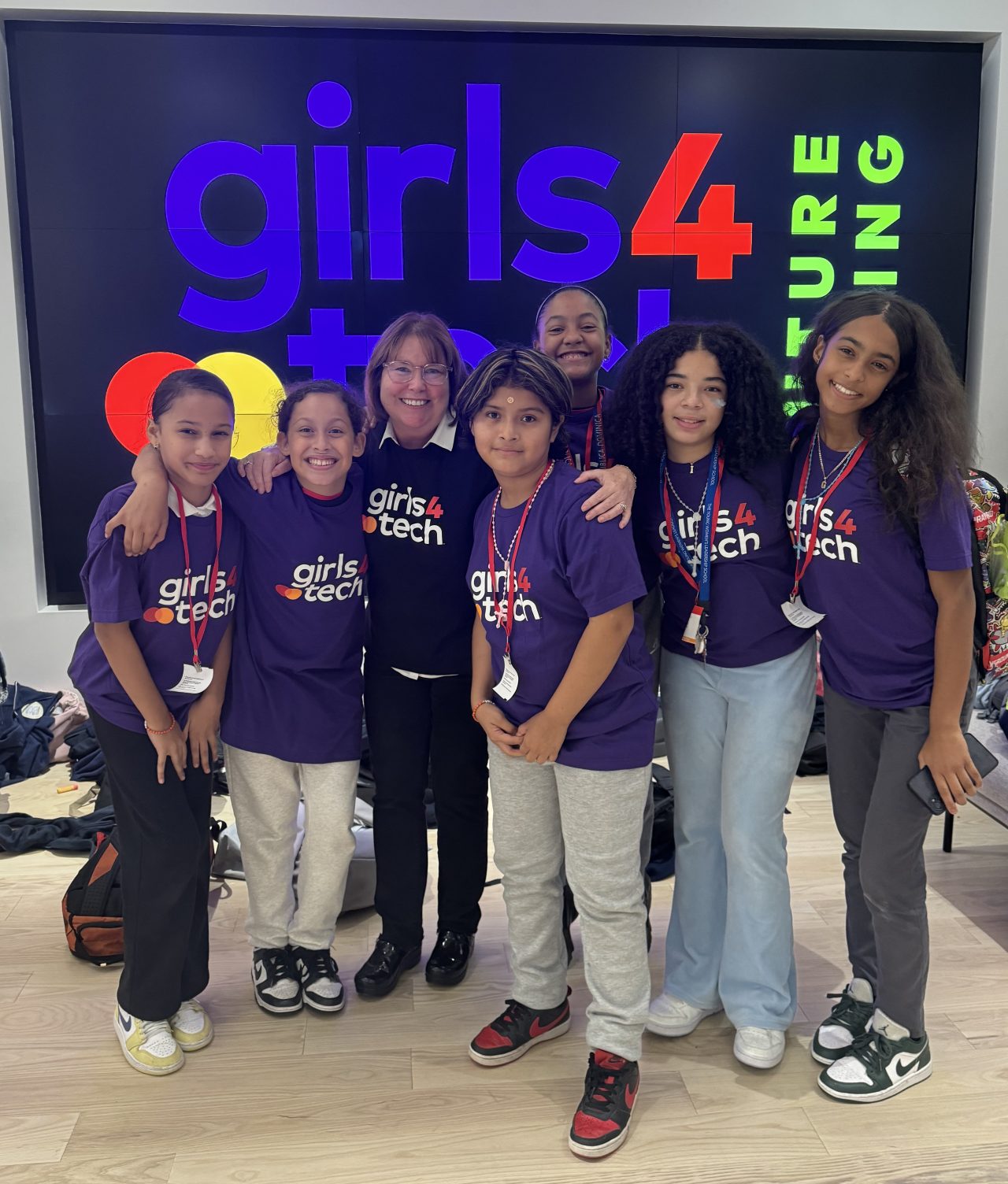 Un grupo de participantes de Girls4Tech con la fundadora Susan Warner vistiendo camisetas de Girls4Tech posando frente al letrero de Girls4Tech Futurecasting.