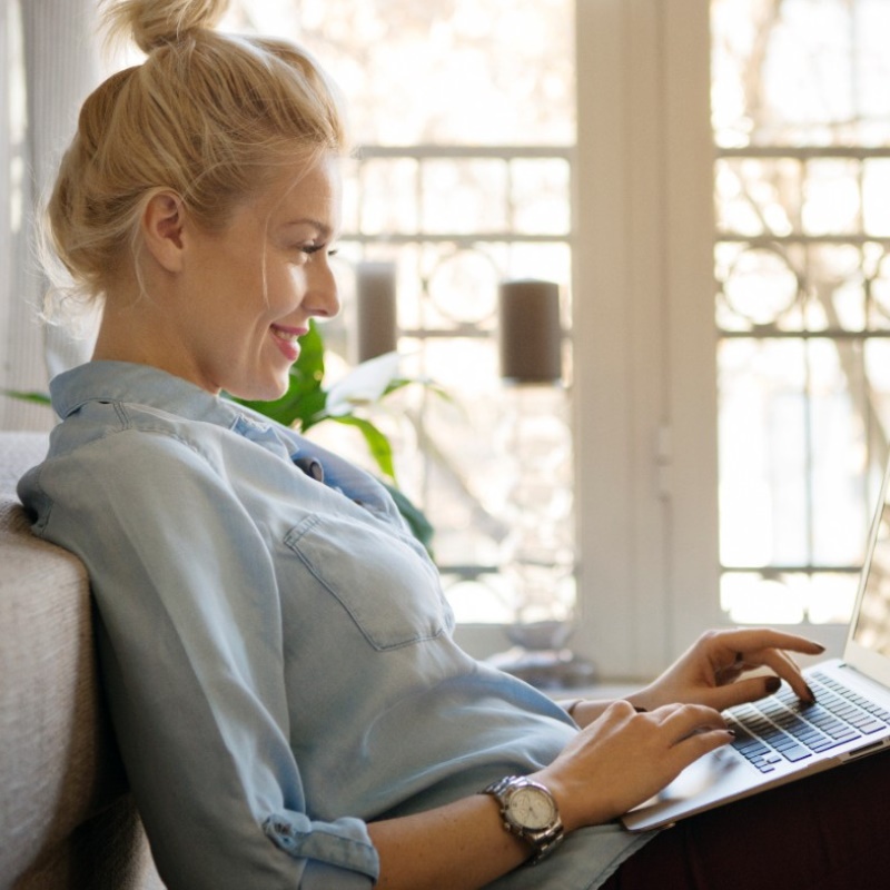 A woman is seated on a couch in her home smiling at her laptop screen.