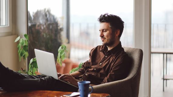 A man sits on a couch looking at his laptop. 