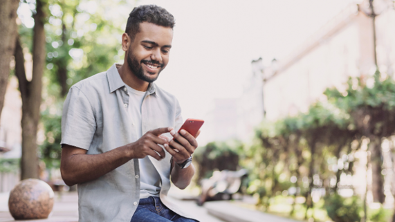 A young man looks at his phone while seated in park lined with palm trees.