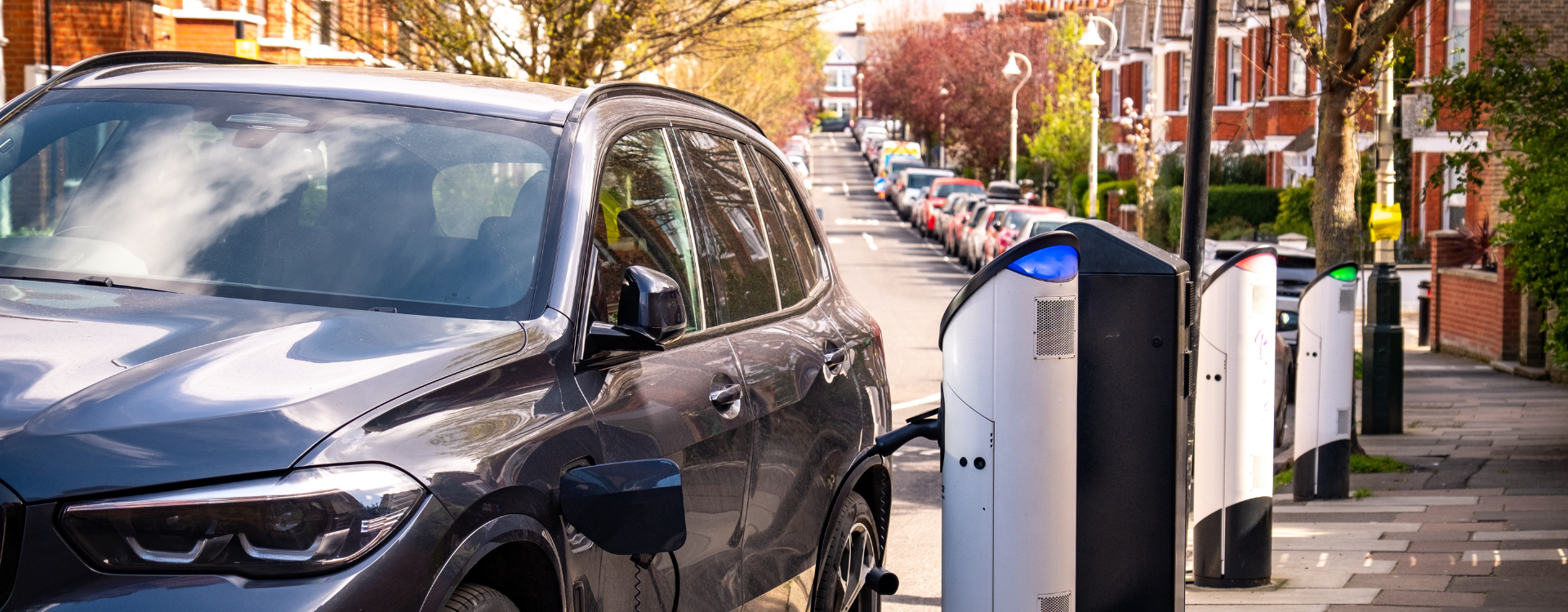 An EV sedan at a curb in a city plugged into an EV charging device.