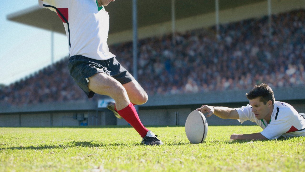 A rugby player holds a ball as another player prepares to kick it in front of stands full of fans.