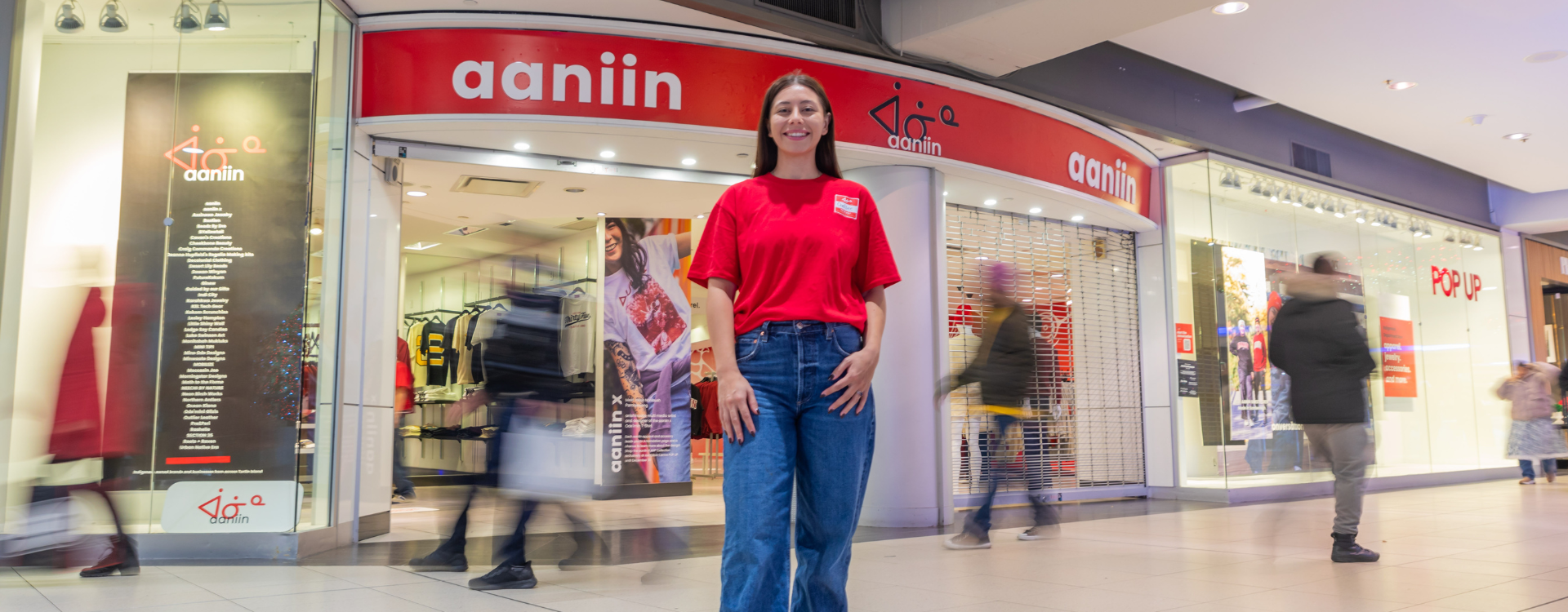 Chelsee Pettit wears a red shirt and jeans in front of her department store, Aaniin, as shoppers walk behind her.