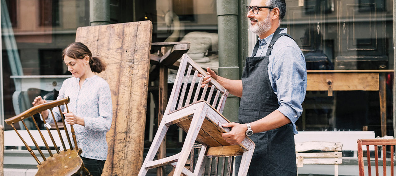To personer utenfor butikk eller verksted, holder stoler de har restaurert eller laget selv.