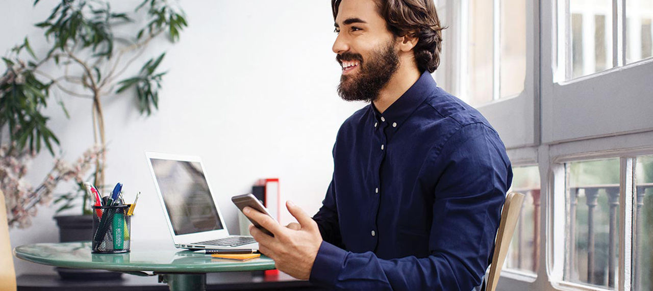En person som smiler mens de holder en mobiltelefon og sitter ved et bord med en bærbar datamaskin.